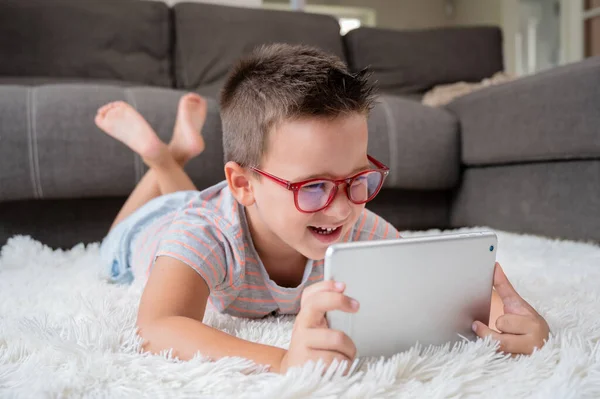 Cute little boy watch cartoons on digital tablet. Kid lies on floor laughing using electronic device. Indoor leisure for children. High quality photography