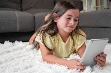 Küçük tatlı kız dijital tablette çizgi film izliyor. Çocuk yerde yatıp elektronik aletle gülüyor. Çocuklar için kapalı bir eğlence. Yüksek kaliteli fotoğrafçılık.