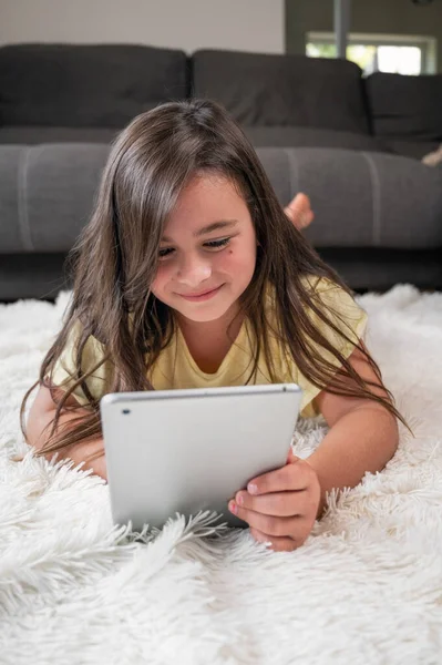 Cute little girl watch cartoons on digital tablet. Kid lies on floor laughing using electronic device. Indoor leisure for children. High quality photography.