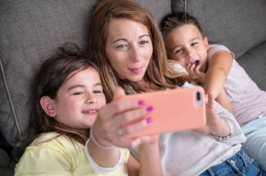 Happy mother with her kids are making a selfie or video call to father or relatives in a sofa. Concept of technology, new generation, family, connection, parenthood. High quality 4k footage