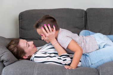 Kanepede uzanmış küçük oğlunu yanağından öpen bir kadın. Anne ve küçük oğlu keyifle evde rahat bir oturma odasında vakit geçiriyorlar. .