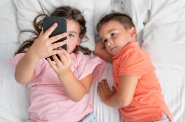 Akıllı telefon kullanan ve evde yatakta yatarken gülümseyen kardeşlerin en iyi görüntüsü. Yüksek kaliteli fotoğrafçılık. Yüksek kalite fotoğraf