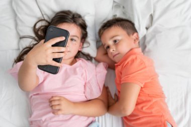 Akıllı telefon kullanan ve evde yatakta yatarken gülümseyen kardeşlerin en iyi görüntüsü. Yüksek kaliteli fotoğrafçılık. Yüksek kalite fotoğraf