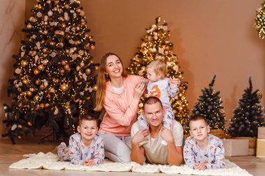 Beş kişilik bir aile Noel ağaçlarının önünde halıda yatıyor.