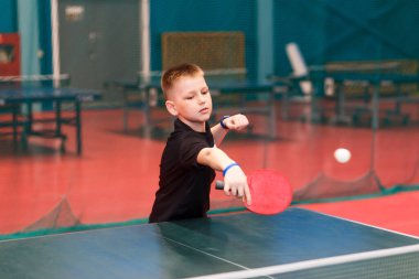 10 yaşında siyah tişörtlü bir çocuk spor salonunda masa tenisi oynuyor.