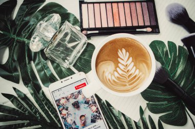 Yazın bir fincan kahve, göz farı paleti ve beyaz ahşap arka planda telefon.