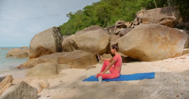 Sportovní žena, která má videohovor, sedí na pláži po cvičení na modré podložce jógy a mluví online, proti tropické přírodě
