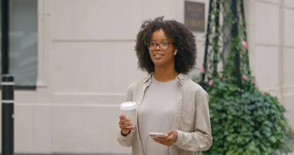 Pretty nice curly haired African American young woman in casual cloth holding cell phone and coffee, having a conversation while enjoying a city walk and looks a way. High quality photo