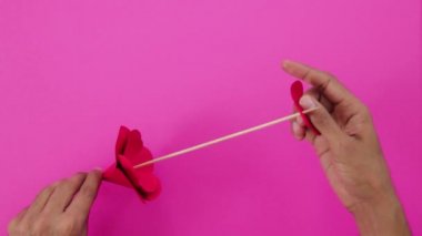 Kartondan kırmızı bir gül yapmak. Yapraklar bir bambu ya da tahta bir çubuğa saplanmış. Kahverengi eller. Yaratıcı el sanatları. Pembe arkaplan.