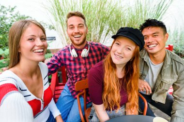 Mutlu kahkahalarla gülen bir grup genç arkadaş açık hava verandasında bir masanın etrafında grup halinde selfie çekiyorlar.