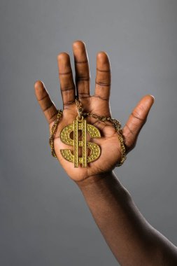 Siyahi bir adam, elinde bir kolyenin üzerinde altın bir sembol tutuyor ve onu parmağında zincir ile avucunun içinde sergiliyor.
