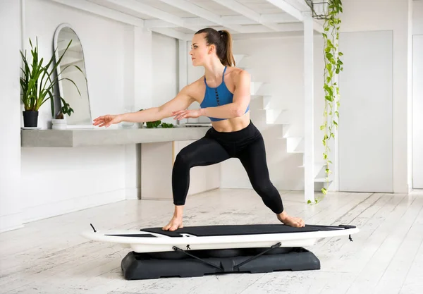 Athletic fit young woman performing a regular surfer pose on a surfset fitness board in a high key gym with potted plants in a health and fitness concept