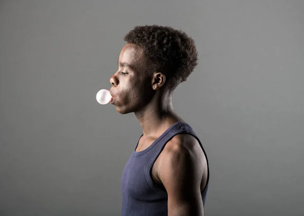 Side view of a trendy sporty young Black man in T-shirt blowing bubbles with chewing gum over a grey studio background in a lifestyle portrait