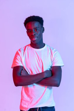 Kendinden emin, siyah saçlı, beyaz tişörtlü, mor arka planda kolları çapraz duran ve kameraya bakan Afro-Amerikan erkek manken.