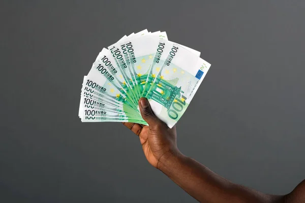 Black hand holding large fan of 100 Euro banknotes with a studio background.