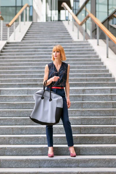 Full body of young fashionable female wearing stylish dark blue pant suit and high heels holding large handbag while standing on stairway near modern urban building in summer day and looking away