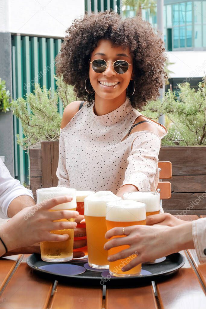 Cheerful black female with curly hair in stylish clothes and sunglasses sitting at table and clinching with glass of beer with friends while spending time on terrace of cafe