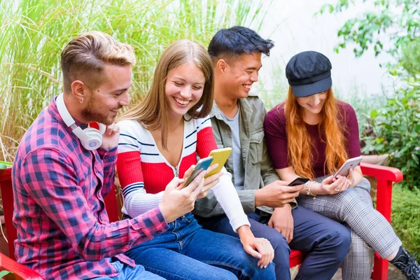 Group of multicultural happy friends using their mobile phones outdoors on a bright red park bench sharing media laughing and joking together in a high key portrait