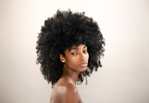 Charming African American female with black Afro hairstyle and bare shoulders looking over shoulder while standing on gray background in light studio