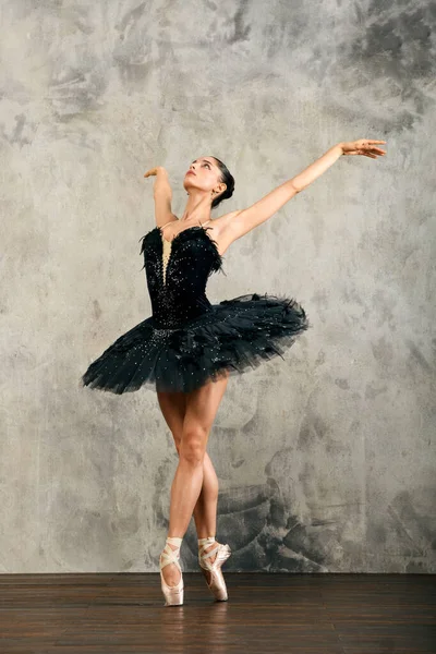 Full body of classical ballet dancer in black tutu standing on tiptoes with raised arms while dancing in light studio