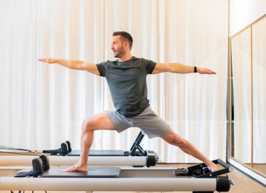 Formda bir adam hamlesini yapıyor ve geriliyor. Savaşçı 2 yoga pozu veriyor. Kalça ve göğüs kaslarını güçlendirmek ve dayanıklılığını arttırmak için pilates yenilikçi yatağı üzerinde duruyor.