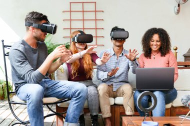 Multiracial men and women in casual clothes with VR helmets interacting with cyberspace while sitting near girlfriend browsing game on laptop on sunny day on patio