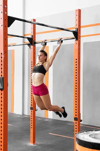 Full body strong female athlete in shorts and bra hanging on pull up bar in arch position while doing kipping exercise during functional workout in gym