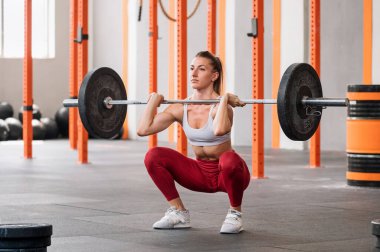Aktif kıyafetli, atkuyruklu, dik bakışlı, halterle ağırlık kaldırma egzersizi yapan, geniş spor salonunda ağırlık kaldırma egzersizi yapan bir sporcu.