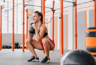 Kararlı bayan sporcu, geniş hafif spor salonunda yoğun antrenman sırasında kafasını yana çevirip ağır kettlebell ile kadeh kaldırıyor.