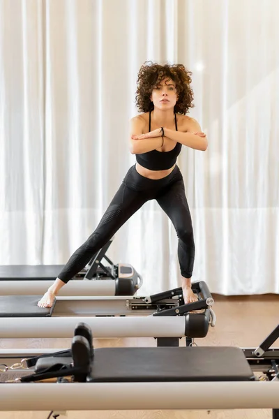 Full body of sportive barefoot female in activewear performing skate pilates pose on reformer bed during training in light gym