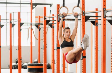 Muscular female athlete in sportswear swinging on gymnastic rings and raising legs during calisthenic workout in sunlit gym