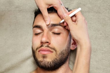 Top view of anonymous female cosmetician using tweezers to pluck eyebrow hair of calm young male client during work in beauty studio