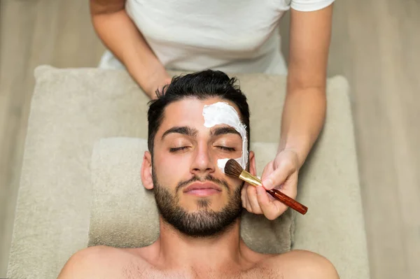 Top view of anonymous woman using soft brush to smear moisturizing mask on face of bearded man with closed eyes during spa session in salon
