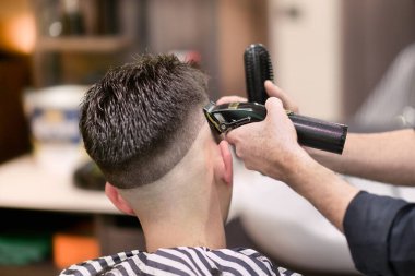 Unrecognizable male barber using electric razor to shave dark hair of man during work in barbershop