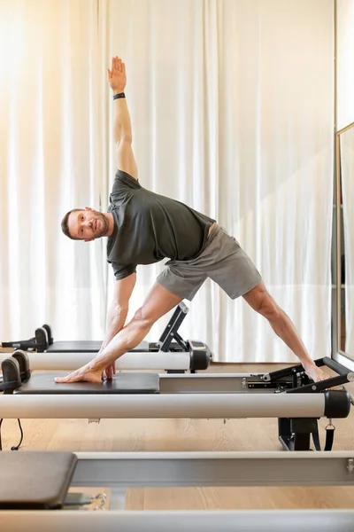 Full body of sportive barefoot male in activewear practicing triangle pose on reformer bed during pilates training in modern gym