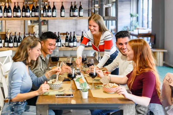Positive diverse friends eating delicious food at table with glasses of red wine during dinner together in modern light restaurant