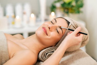 Tarlada tanınmayan bir güzellik uzmanı, kaşlarına pudra sürerken mükemmel derisi havluya sarılmış ve kaplıca ameliyatından sonra masaya uzanmış genç bir bayan müşteri.