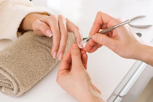 From above nail technician cutting cuticle on finger of woman holding hand on rolled up towel on white table in spa salon