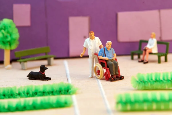 身穿制服的塑料迷你男护士在临终关怀大楼前帮助坐轮椅的老年病人 — 图库照片
