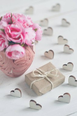 Kraft kağıdına sarılı güzel kompozisyon ve beyaz ahşap arka planda pembe vazoda narin güller. Küçük tahta kalplerle kompozisyonu süsleyin..