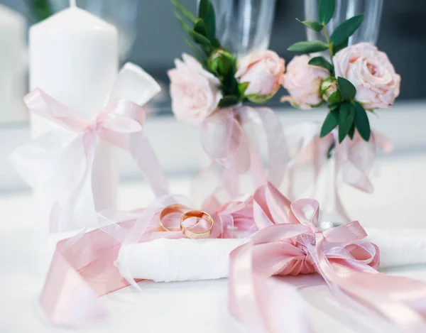 Two gold wedding rings lie on the table next to the wedding accessories. Wedding rings lie on the table near the candles with pink ribbons and glasses decorated with pink flowers. Blurred background.