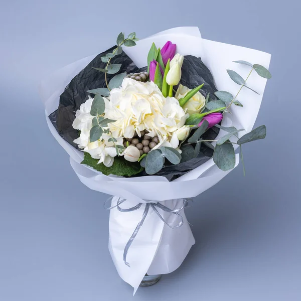 Delicate romantic bouquet of flowers wrapped in white paper. Festively packaged bouquet of tulips, white hydrangea and eucalyptus twigs on a blue background. Floristics concept.