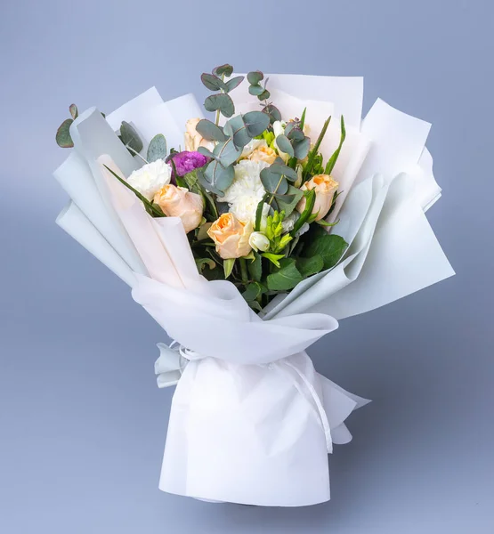 Delicate romantic bouquet of flowers wrapped in white paper. Beautifully packaged floral arrangement with pale pink roses, white peonies and eucalyptus twigs on a blue background.