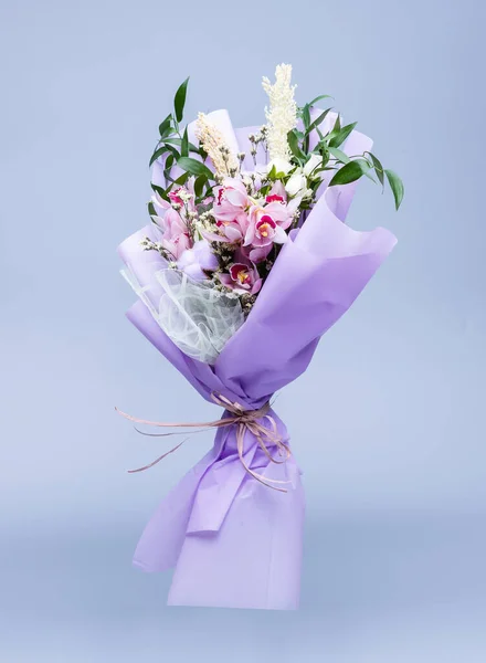 Bouquet of fresh natural flowers wrapped in floral tracing paper stands in a vase on a blue background. Bouquet in pink style composed of irises, other different flowers and twigs of cotton.