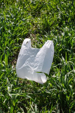 White small plastic bag lies on green grass in park or in field under sun. Concept of ecology, waste plastic bags, nature conservation and cleaning earth from plastic.