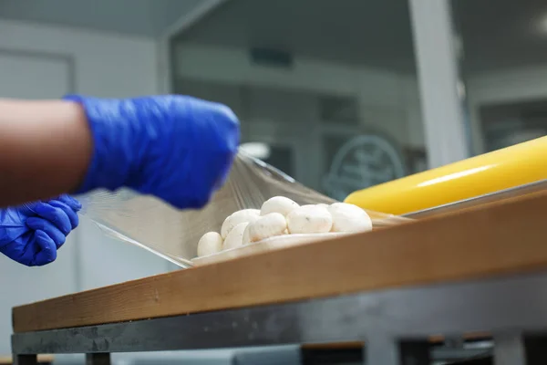 Worker wraps mushrooms lying in foam dish in stretch film. Close up of man\'s hands in disposable gloves making portions of mushrooms wrapping them in cling film.