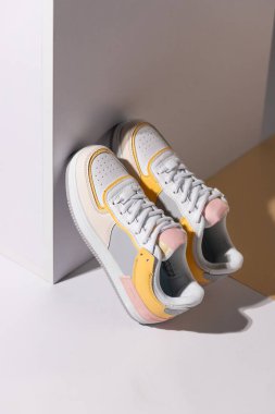 Close up of pair of colored sneakers standing leaning against white wall in studio. Women's white sneakers with yellow and pink inserts stand, leaning on cube. Spring and summer fashion concept.