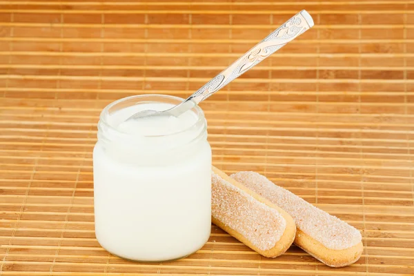 Frasco con yogur casero y cuchara, por una galleta en una estera de paja — Foto de Stock