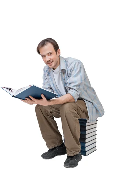 Junger Mann sitzt auf Bücherstapel, hält Buch in der Hand und blickt in die Kamera — Stockfoto
