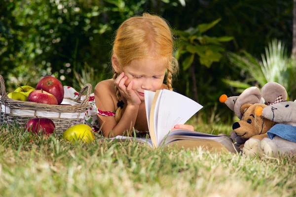 Menina lendo um livro com brinquedos ao ar livre — Fotografia de Stock